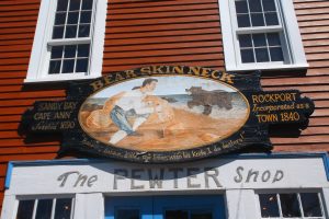 A colorful sign reading "Bear Skin Neck" with a painted scene of a man confronting a bear, flanked by text "SANDY BAY CAPE ANN Settled 1690" on the left and "ROCKPORT Incorporated as a TOWN 1840" on the right, mounted on an orange, clapboard building above "The PEWTER Shop" sign.