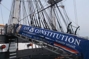 A banner reading "USS Constitution Oldest Commissioned Warship Afloat 'Old Ironsides'" displayed on the side of an historic naval ship with masts and rigging in the background.