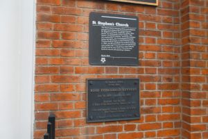 Two commemorative plaques mounted on a brick wall, with the upper plaque describing the history of St. Stephen's Church and the lower plaque dedicated to Rose Fitzgerald Kennedy.