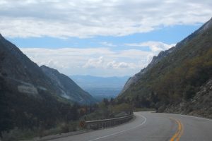 A winding mountain road with yellow lines and a guardrail on the right, with a sprawling valley and distant mountains under a partly cloudy sky.