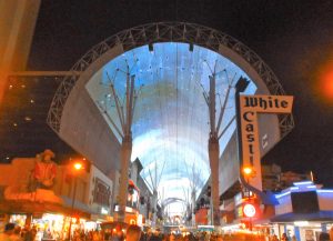 Alt text: A vibrant street scene at night under a large illuminated canopy with a neon sign reading "White Castle" and a crowd of people along the walkway.