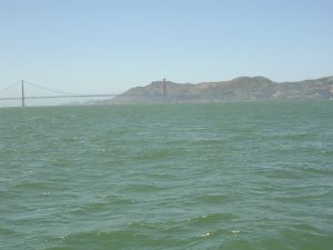 View of a bridge partially obscured by hazy conditions in the distance, with calm green waters in the foreground and hills in the background under a clear sky.