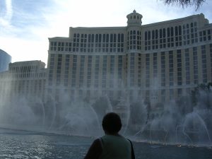 A person is observing a large-scale water fountain show in front of the Bellagio Hotel and Casino during daylight, with the fountain's spray partially obscuring the view of the building.