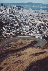 Aerial view of a winding road on a hillside overlooking a dense urban area with skyscrapers in the distance and a body of water beyond the cityscape.