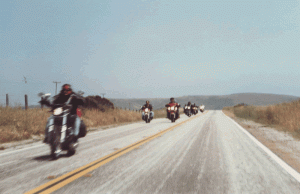 A group of motorcyclists riding in formation down a two-lane highway with a clear sky and open fields on either side.