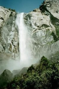 A towering waterfall cascading down a rocky cliff face, surrounded by green vegetation under a blue sky.