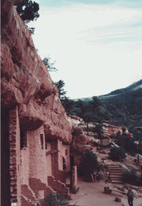 Cliff dwellings built into the side of a reddish-brown rock face with steps leading up to the entrance and trees in the background.