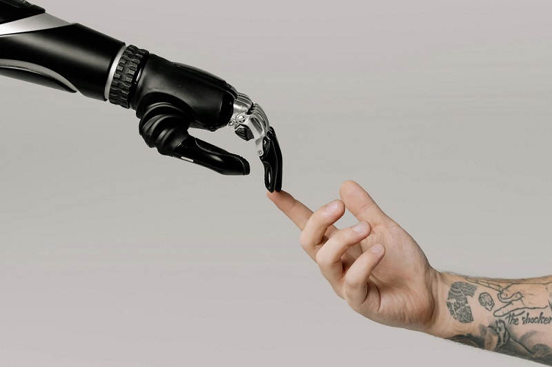 A robotic hand with intricate details touching a human hand with a visible tattoo, symbolizing the connection between humans and technology.