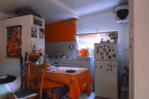 A compact kitchenette featuring a dining table, a refrigerator decorated with magnets, and orange wall accents, showcasing a warm and inviting atmosphere.