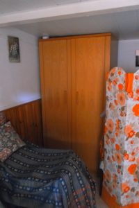 A small bedroom featuring a wooden wardrobe, a bed covered with a patterned blanket, and a floral room divider.