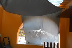 A shaded area featuring a gray fabric covering and an arched window showcasing vibrant blooms outside, located in a sunny setting.
