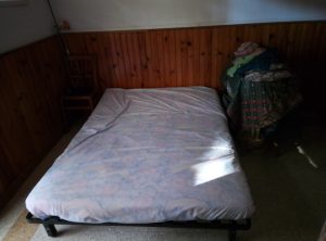 A simple bed with a light-colored mattress in a room accented by wooden walls and a chair nearby, along with a stack of clothes on a small table.