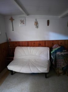 A white futon sofa in a rustic room featuring wooden walls, decorated with various wall hangings and a stack of colorful clothes on a nearby chair.