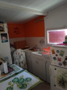 A small kitchen featuring an orange accent wall, a white sink, and a variety of kitchen decor, including a colorful tablecloth and fridge magnets.