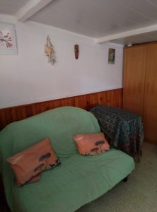 A comfortable green sofa with decorative pillows against a wooden wall, featuring wall art and a corner cabinet draped with a patterned cloth.
